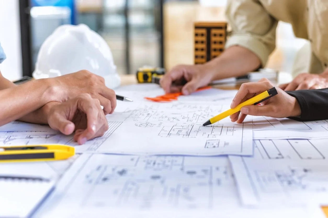 Hands marking up blueprints on a table with a hard hat, tape measure, and pencils nearby