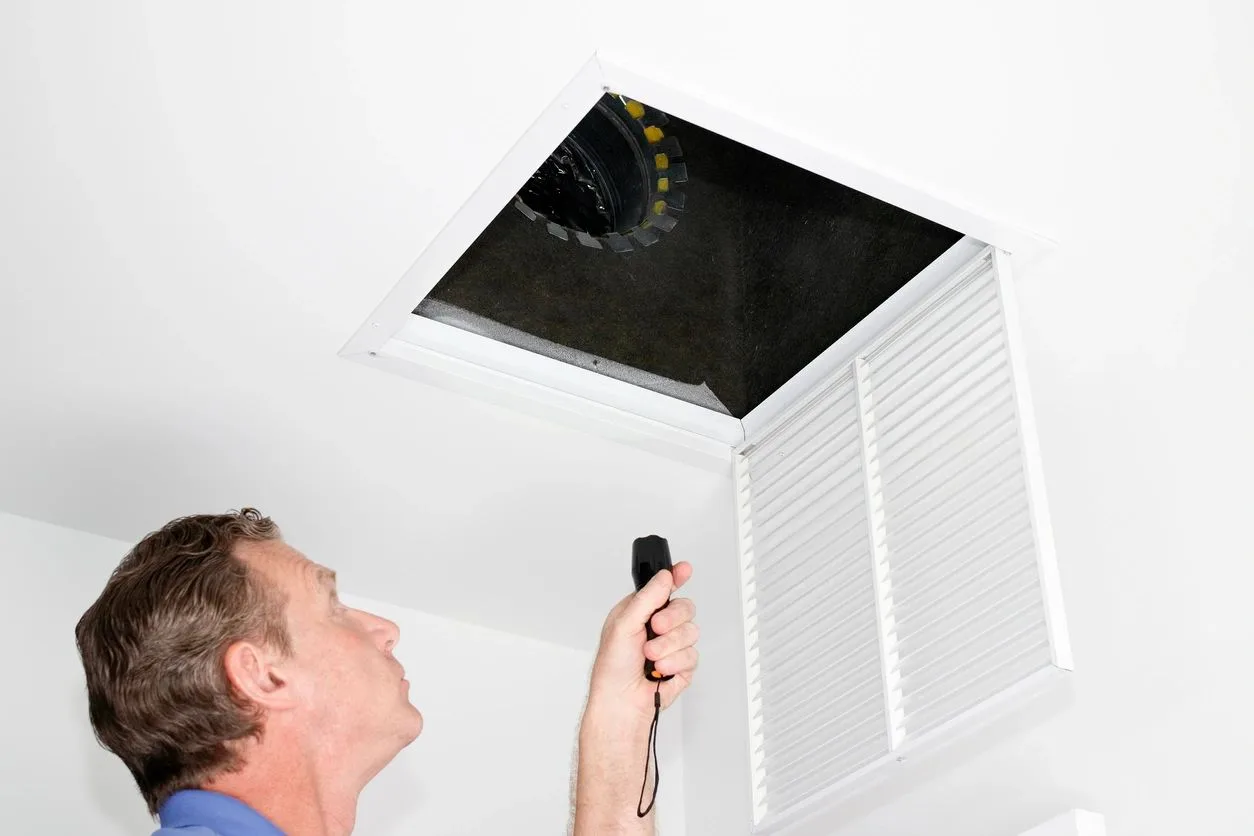 Inspector using a flashlight to examine an attic access opening and ductwork