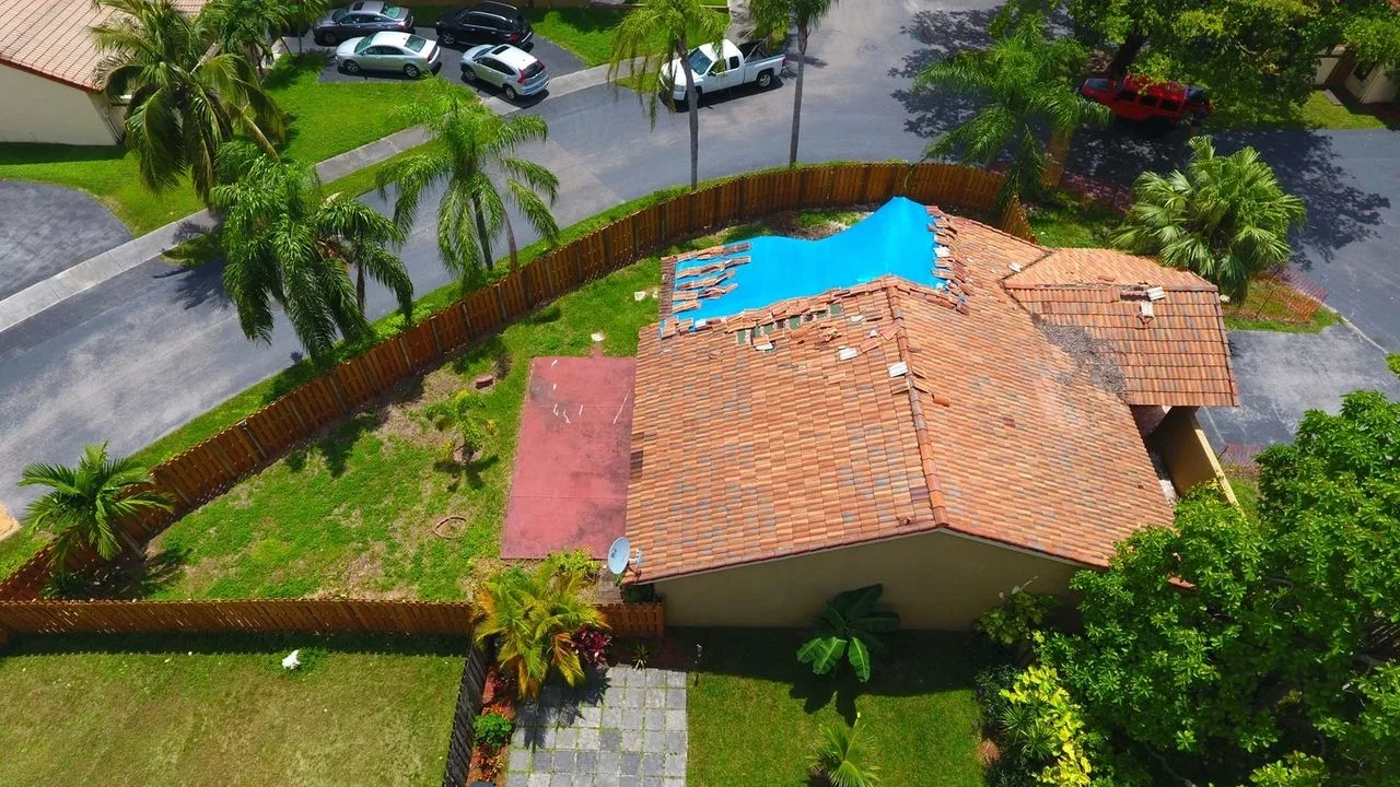 Aerial view of storm-damaged roof tiles and a blue tarp over the pool after a hurricane