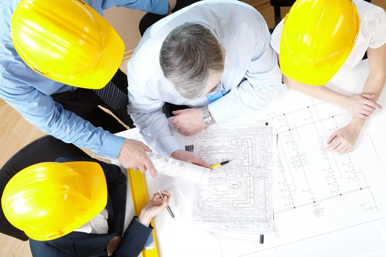 Team in yellow hard hats gathered around a table reviewing blueprints from above